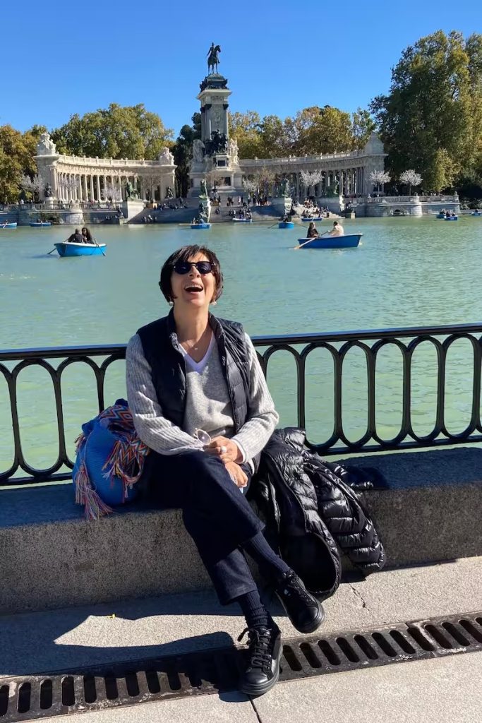 Martha Escamilla laughing, sitting by the lake in the Retiro in Madrid, with the Alfonso XII monument behind her