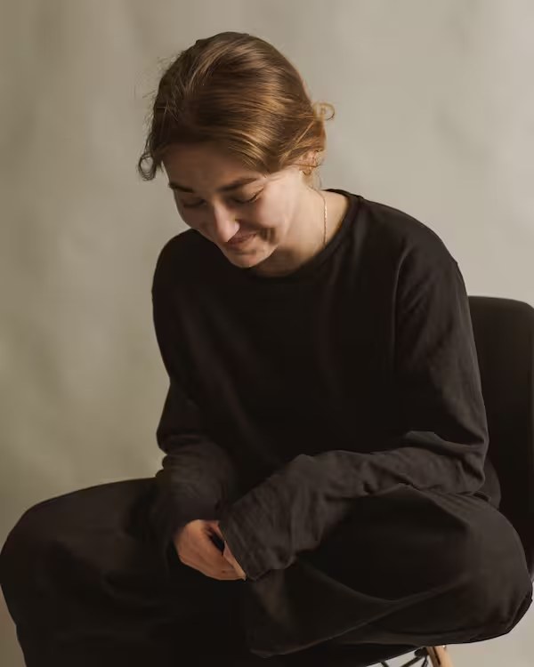 A woman sitting, eyes lowered, with a quiet smile