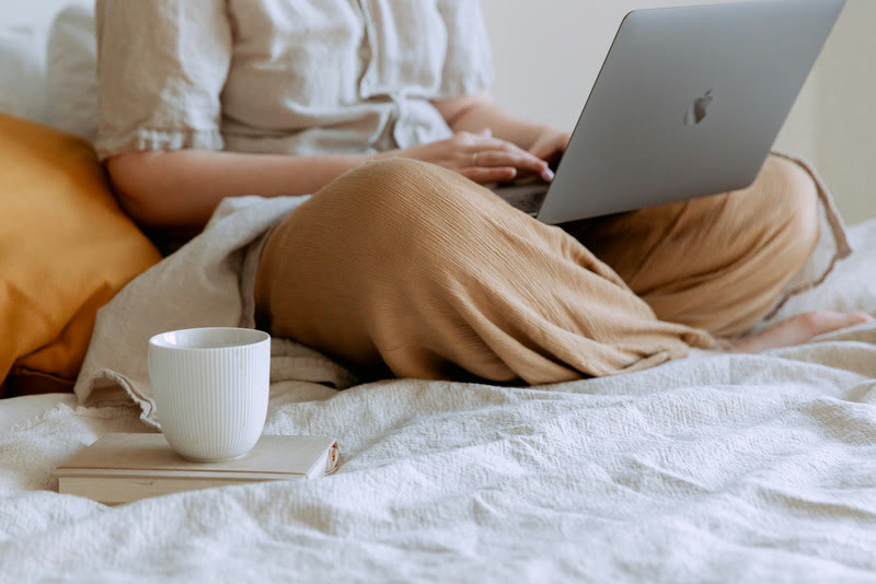 A woman working on her laptop in bed, with a cup and a book